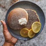 black hand holding blue plate with two lemon ricotta pancakes garnished with lemons and powdered sugar