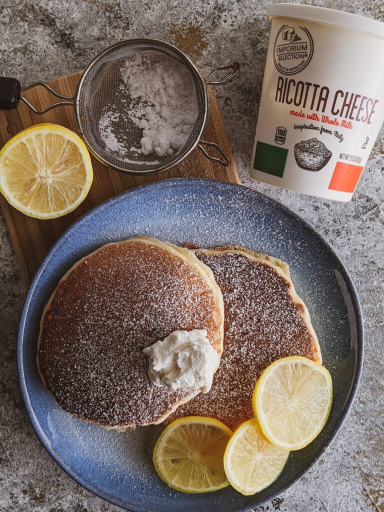 two lemon ricotta pancakes on a blue plate garnished with lemons and powedered sugar
